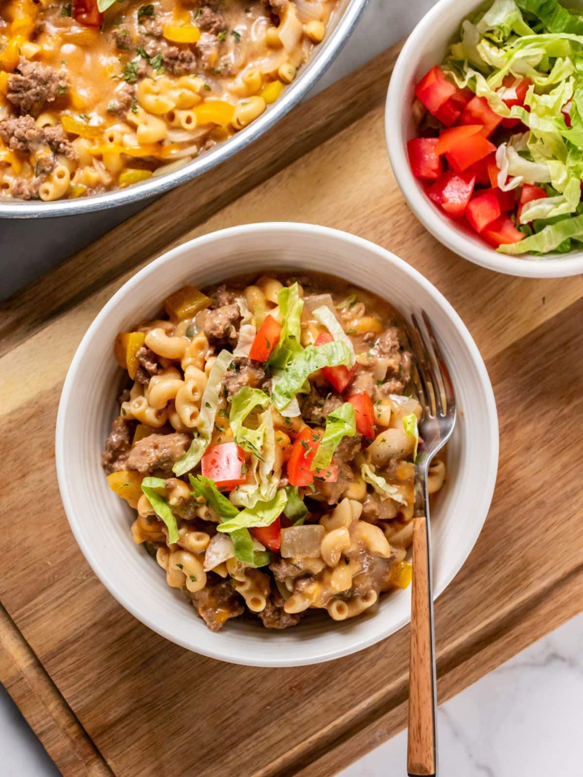 A bowl of cheeseburger pasta with a fork and a small bowl of lettuce and tomato to the side and the casserole dish in the background.