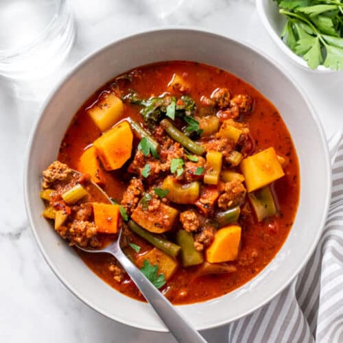 picture of a white bowl with a spoon and tomato-based soup with potato and ground beef