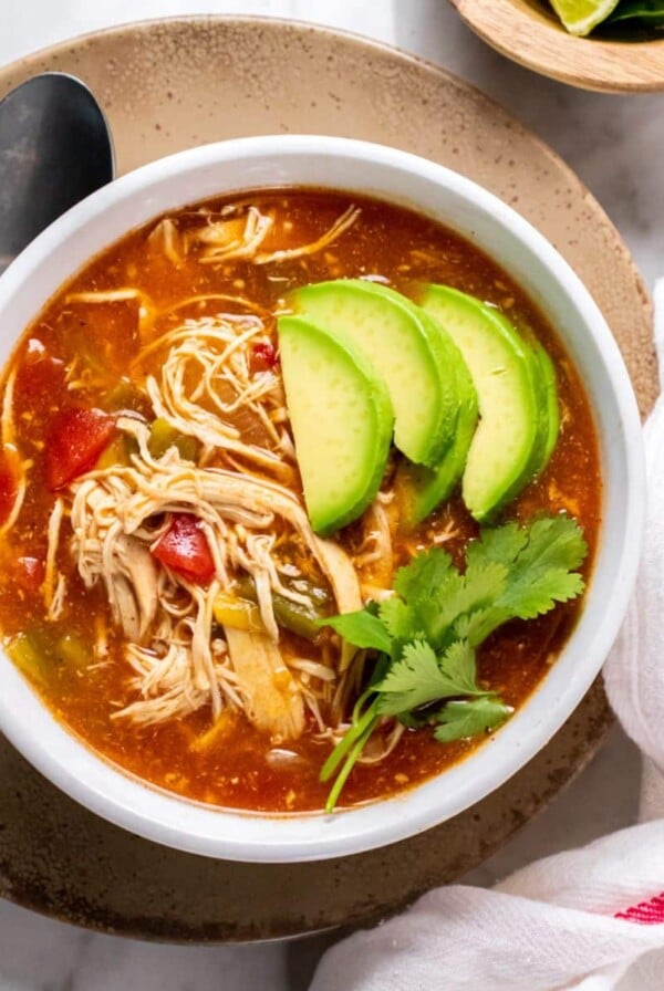 A bowl of healthy chicken enchilada soup on the table topped with slices of avocado and cilantro.