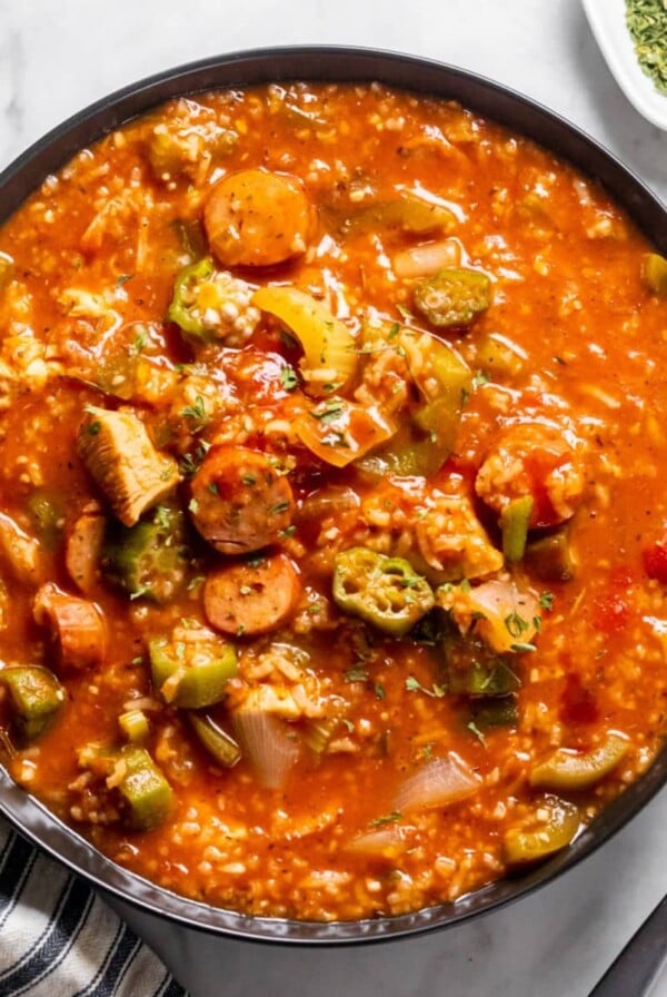 A bowl of crockpot gumbo on the table with some seasoning, a spoon, and a napkin next to the bowl.