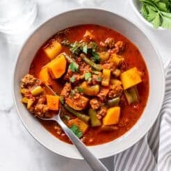 A bowl of healthy hamburger soup on the table with a spoon resting in the bowl and a napkin and bowl of parsley to the side.