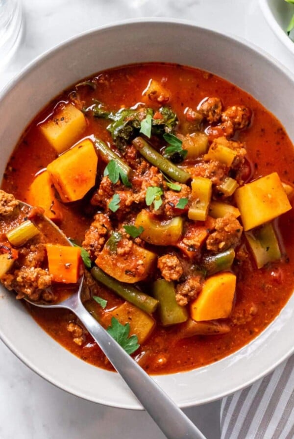 A bowl of healthy hamburger soup on the table with a spoon resting in the bowl and a napkin and bowl of parsley to the side.