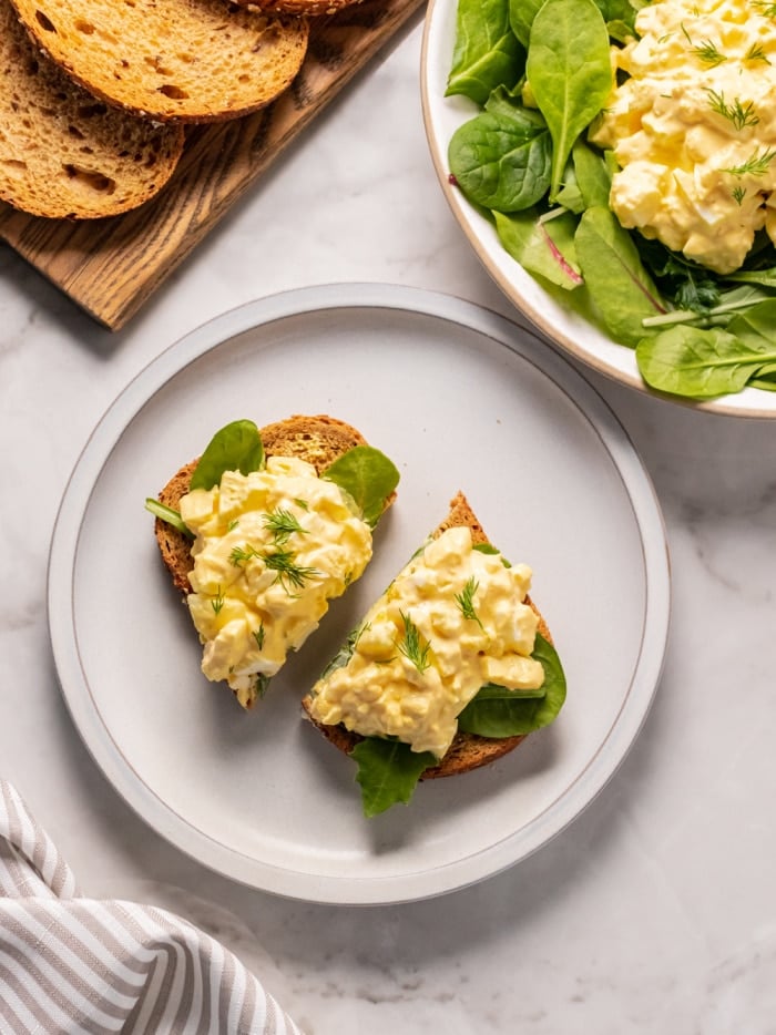 egg salad on two slices of toast on a white plate on the counter