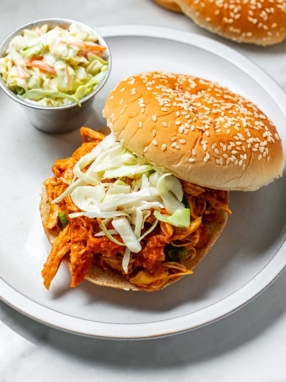 Shredded chicken sloppy joe on a hamburger bun with coleslaw and. a seeded bun tilted to the side.