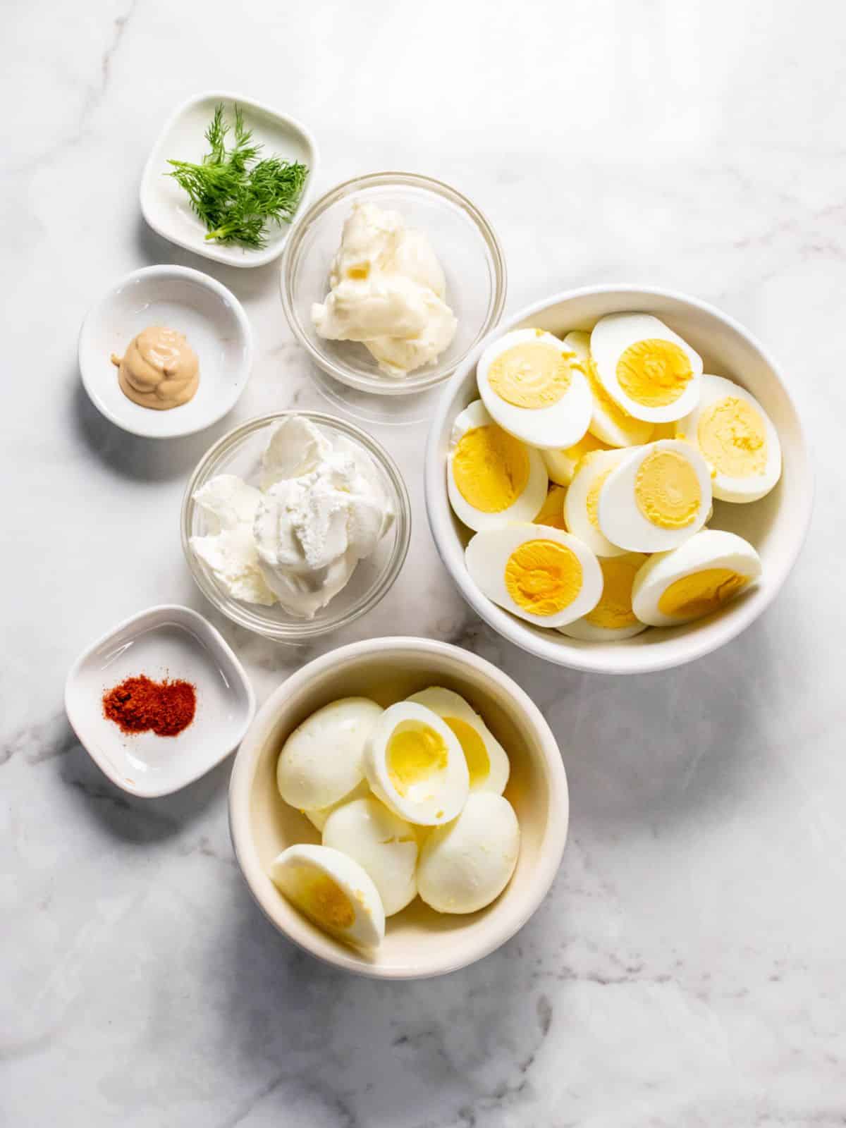 Ingredients to make healthy egg salad in white bowls on a marble surface.