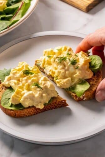 High protein egg salad on a piece of bread with a hand reaching to grab a half.