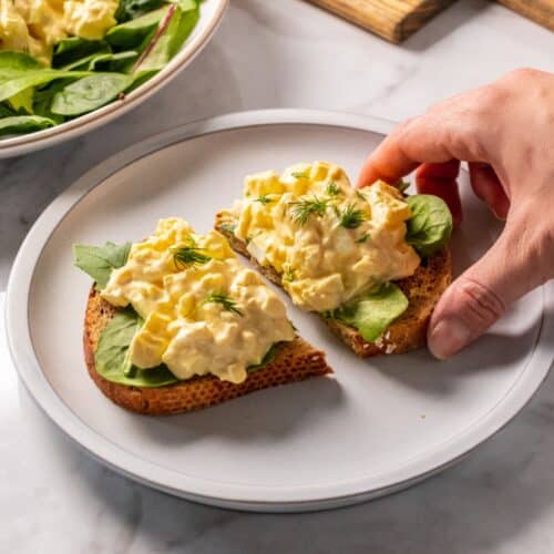 High protein egg salad on a piece of bread with a hand reaching to grab a half.