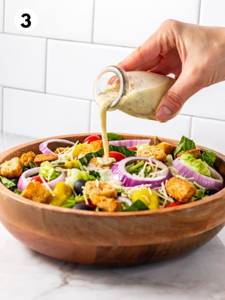A hand pouring a small jar of dressing over a homemade Olive Garden salad.