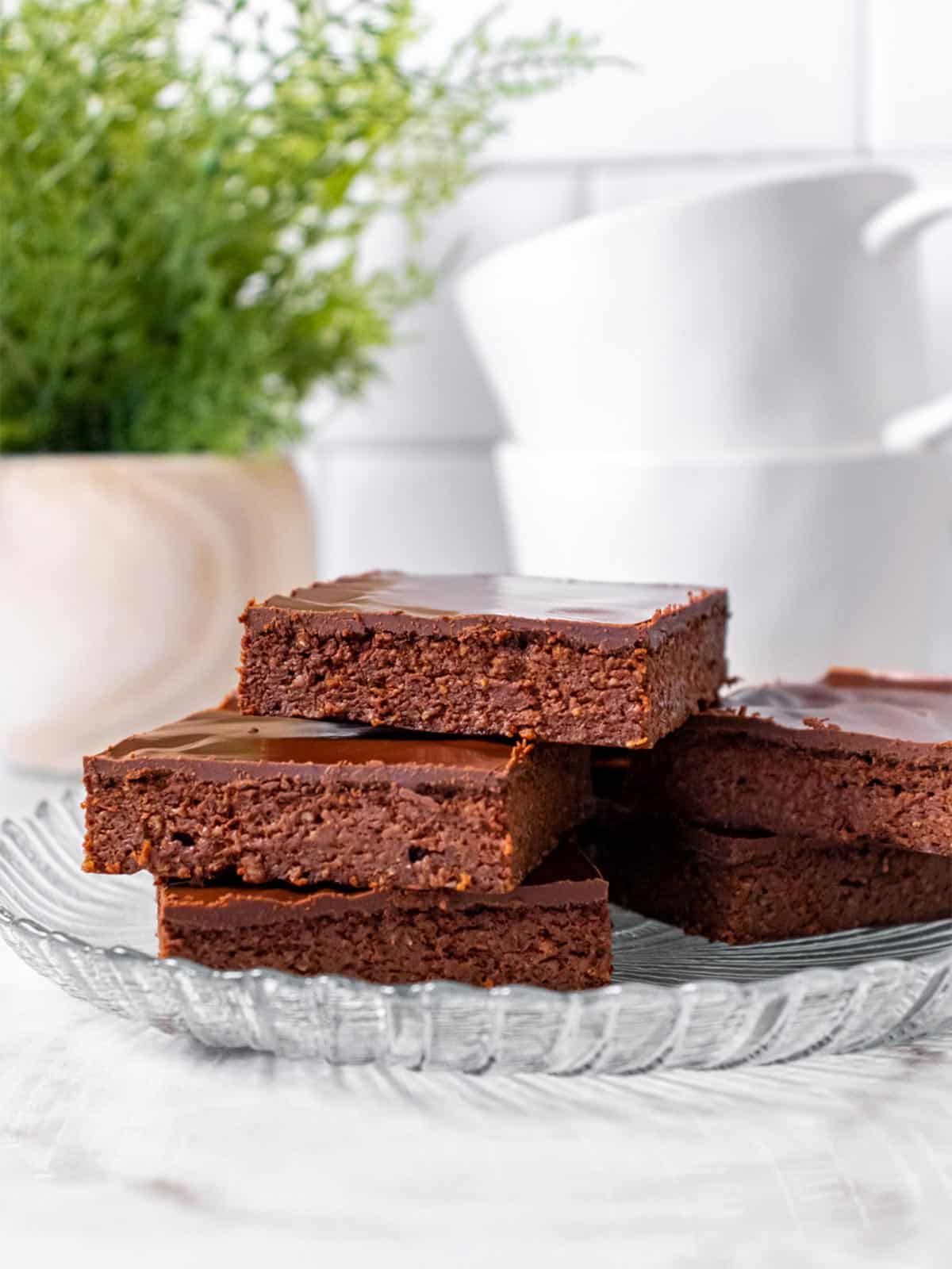 A stack of greek yogurt brownies on a clear plate with white coffee cups in the background.