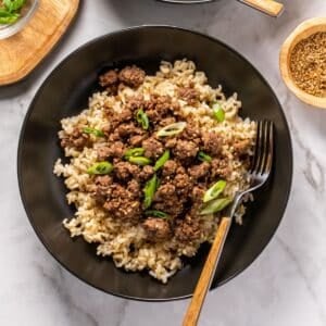 Korean beef bowl on the table with a fork resting on the side and small bowls of toppings around the bowl.