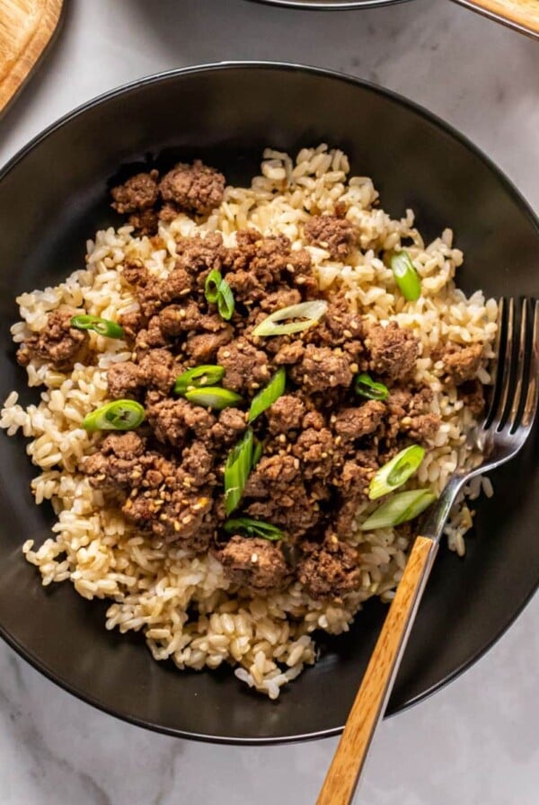 Korean beef bowl on the table with a fork resting on the side and small bowls of toppings around the bowl.