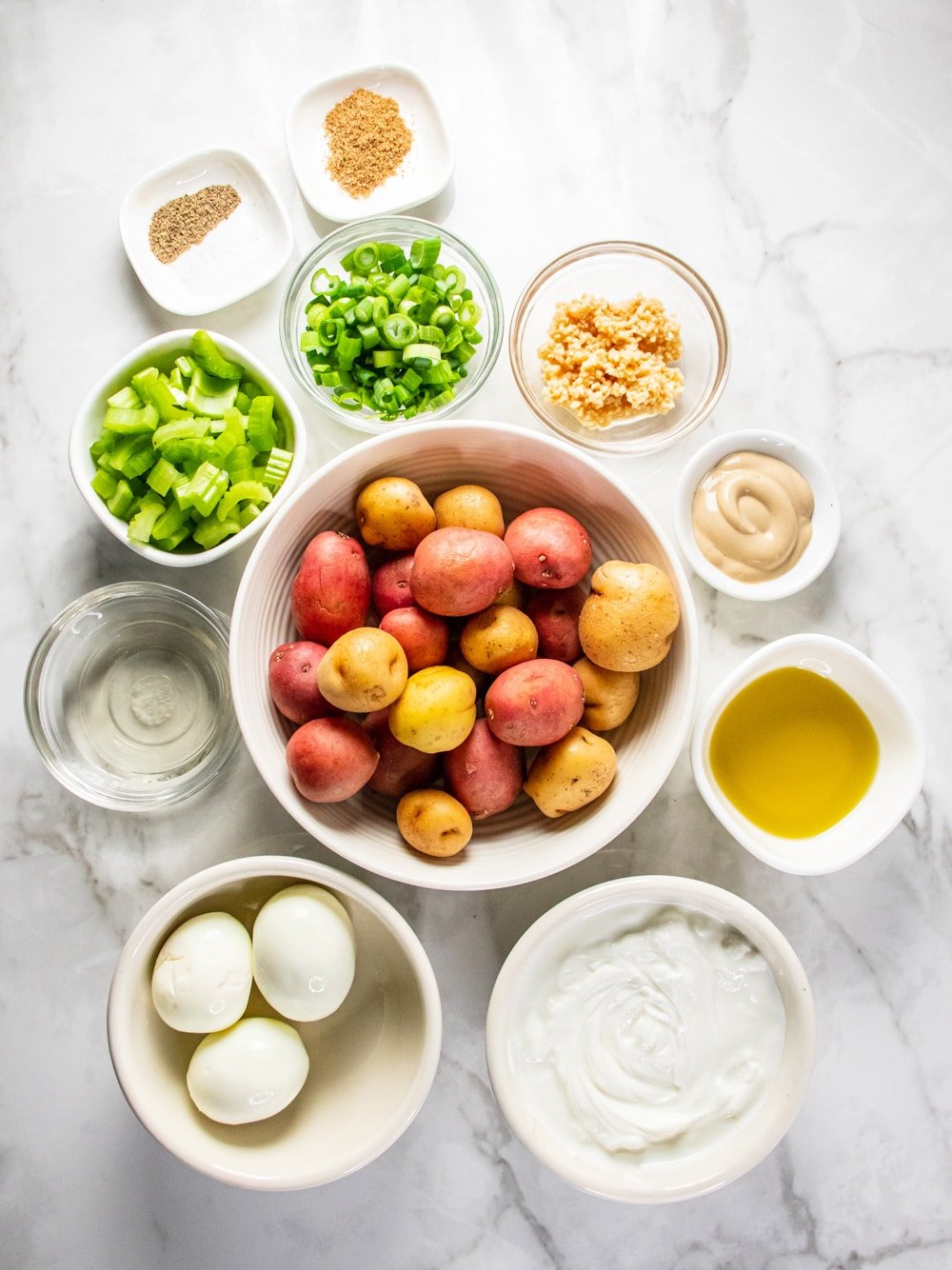 potatoes in a white bowl with ingredients for potato salad in bowls around it