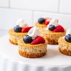 Mini protein cheesecake bites are on a white platter and garnished with whipped cream and fresh fruit.