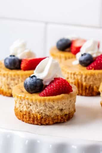 Mini protein cheesecake bites are on a white platter and garnished with whipped cream and fresh fruit.