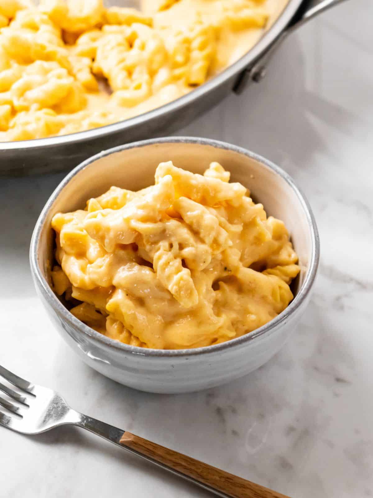 A bowl of high protein mac and cheese on the table in front of a skillet of more. 