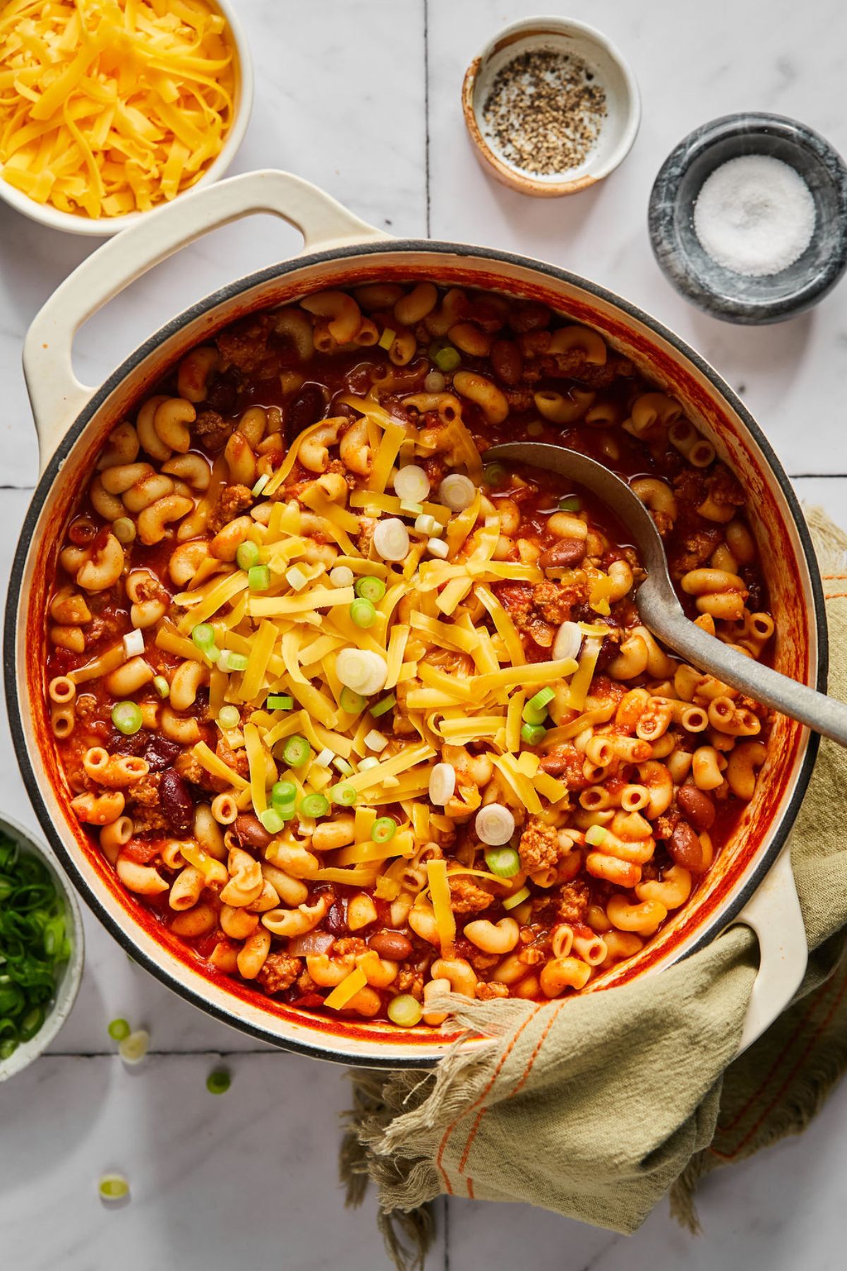 A pot of healthy chili mac on the table with a serving spoon dipping into the dish and cheese and green onions sprinkled over the top.