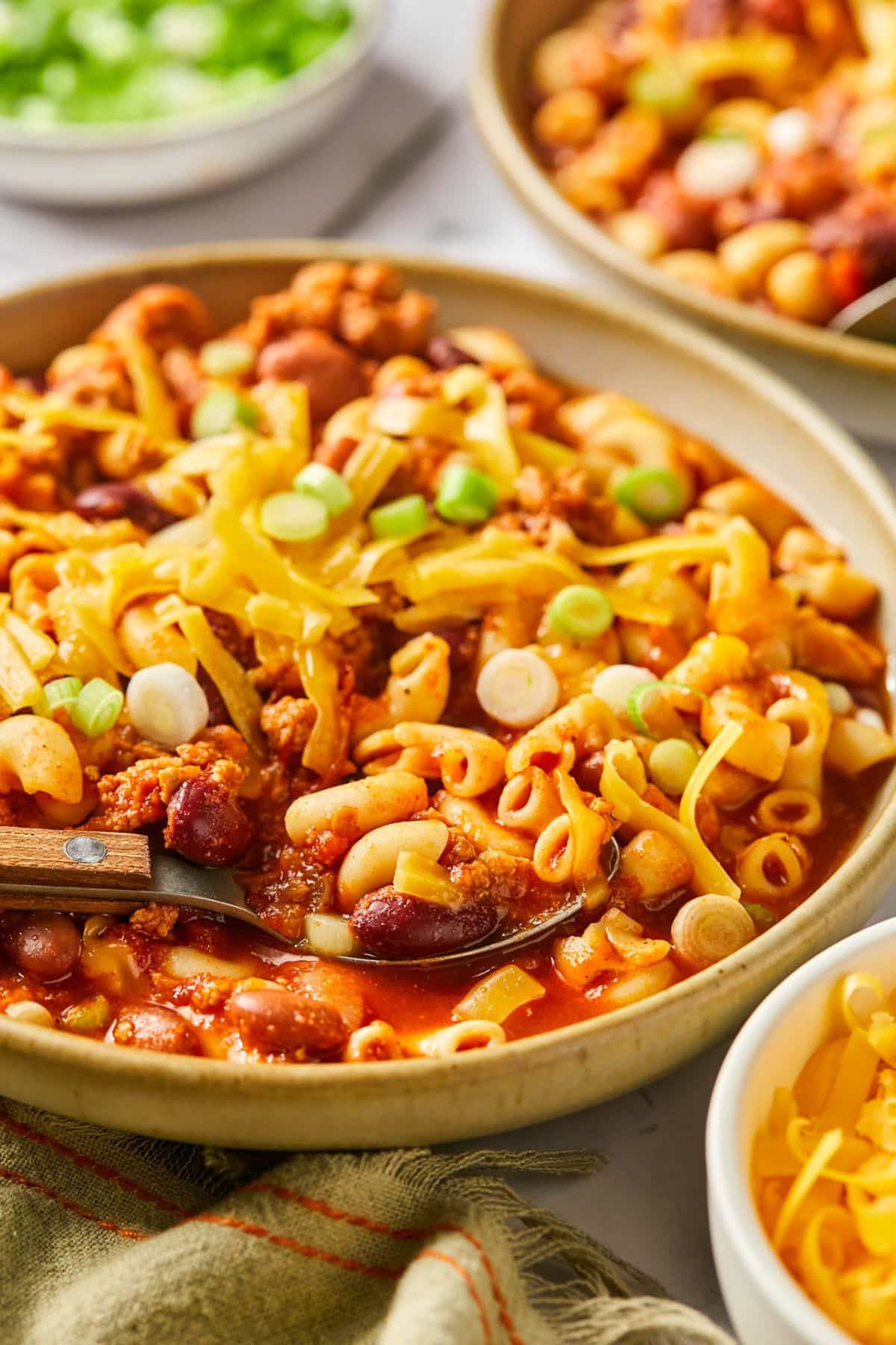 A bowl of turkey chili mac on the table with a spoon in the bowl and more dishes around the bowl of toppings and more chili mac.