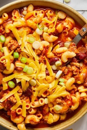 A bowl of healthy chili mac with a spoon in the bowl and cheese and green onions sprinkled on top.