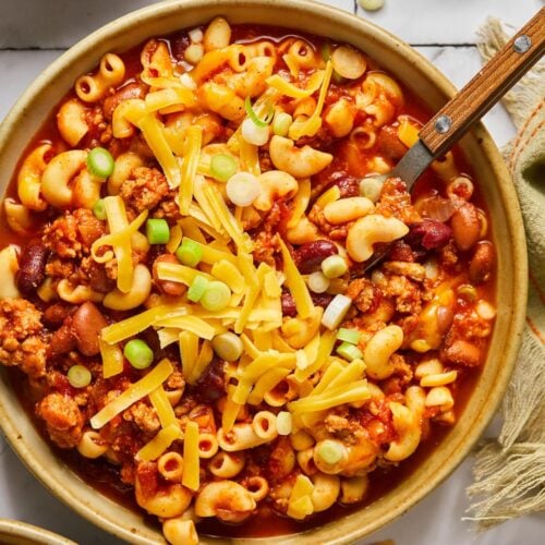 A bowl of healthy chili mac with a spoon in the bowl and cheese and green onions sprinkled on top.