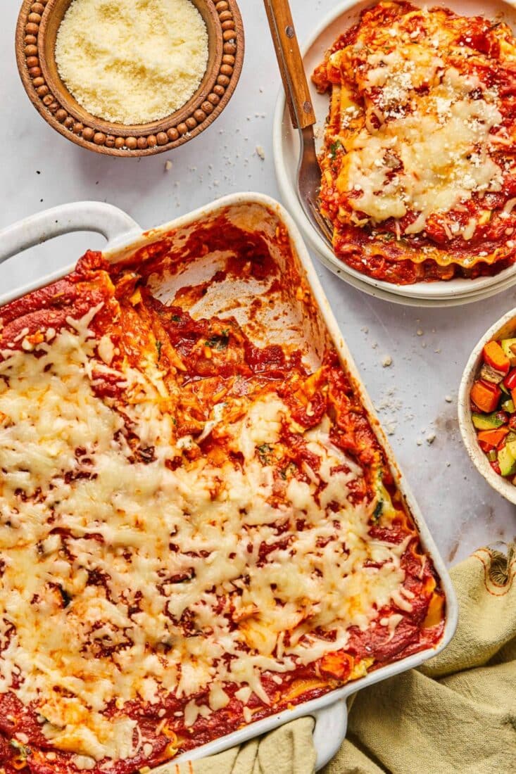 A casserole dish of high protein vegetarian lasagna on the table with plates and bowls of cheese to the side.