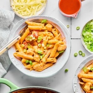 A bowl of buffalo chicken pasta next to a bowl of shredded cheese and buffalo sauce.