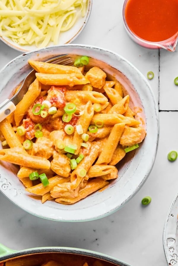 A bowl of buffalo chicken pasta next to a bowl of shredded cheese and buffalo sauce.
