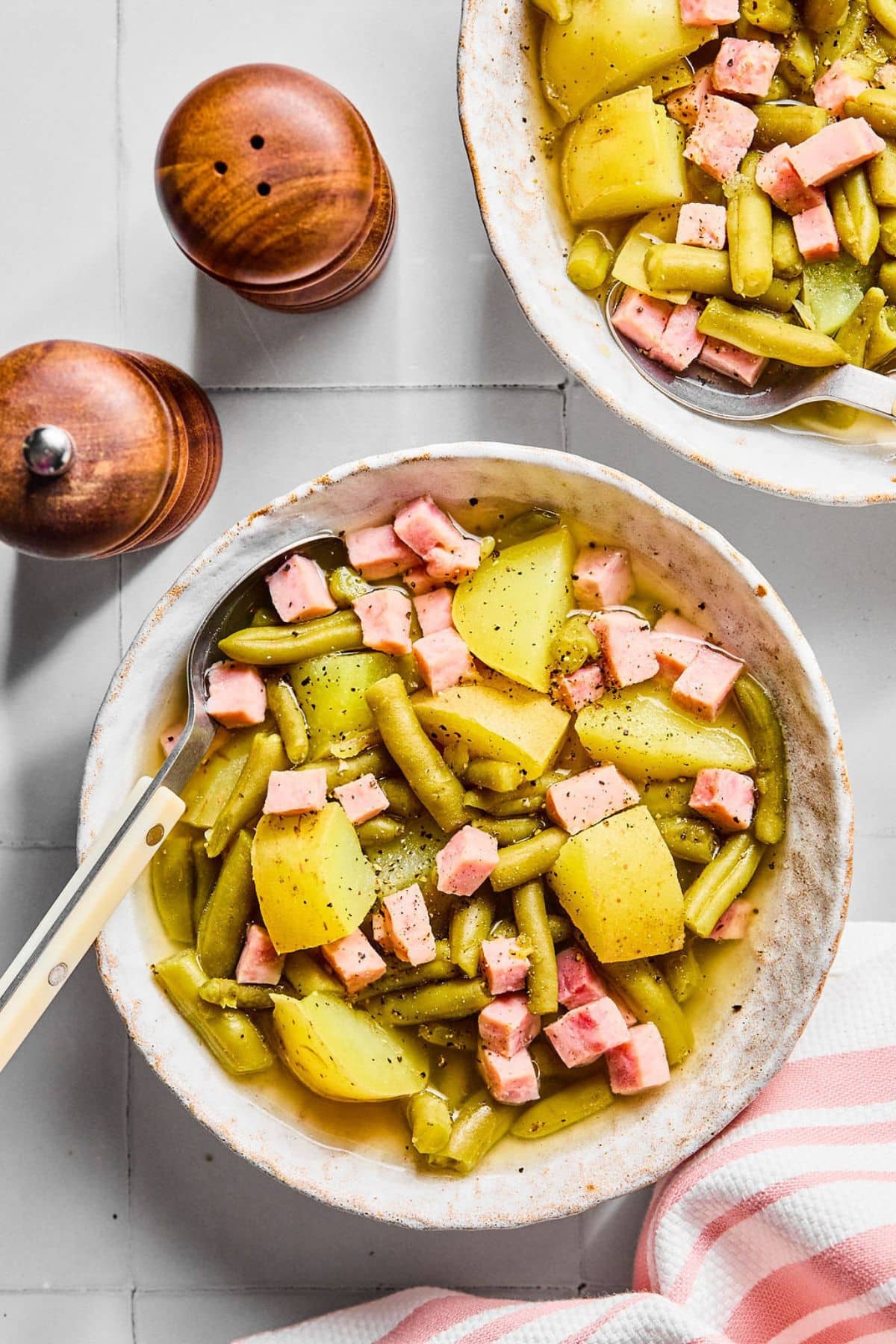 A bowl with ham green beans and potatoes with a spoon on the side.