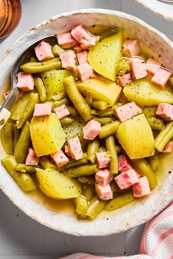 A bowl filled with ham, green beans, and potatoes and a utensil resting to the side.