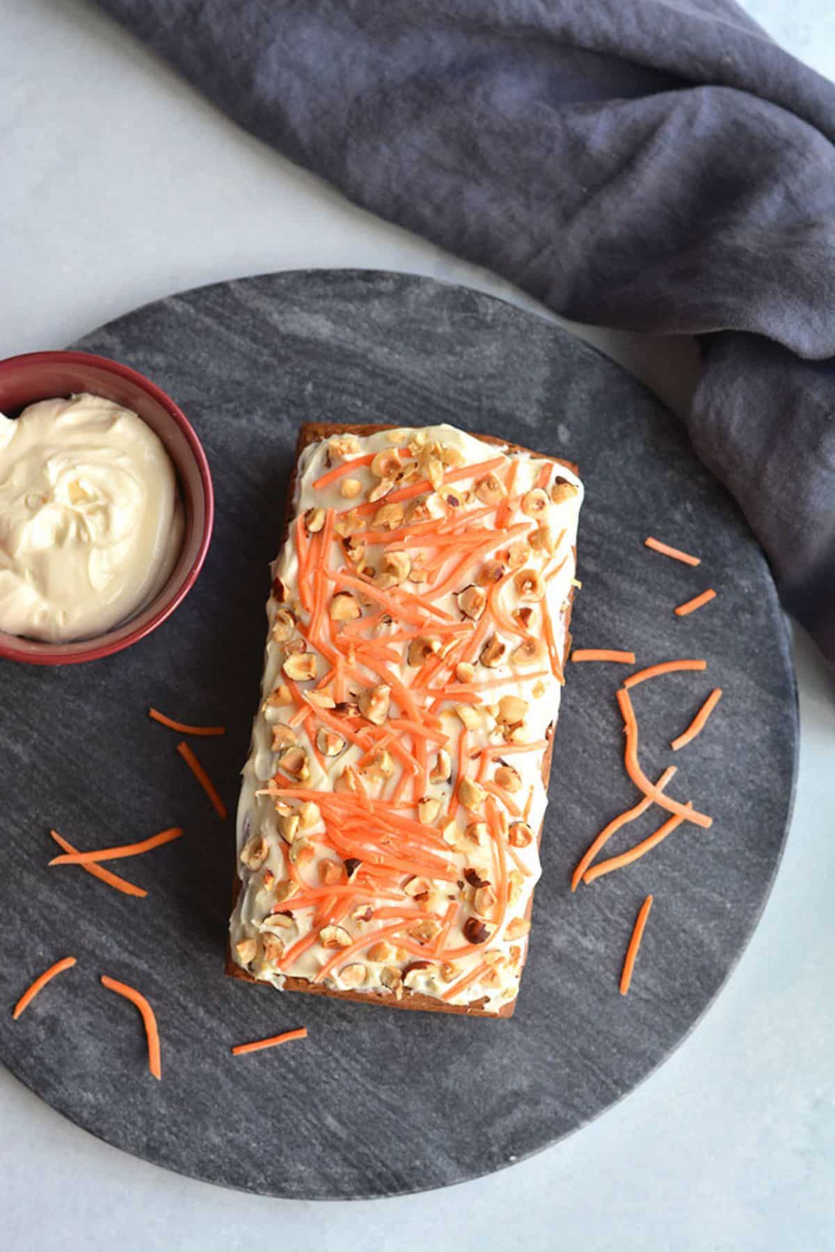 A loaf of banana carrot bread on a round tray next to a bowl of icing and shredded carrots scattered around.