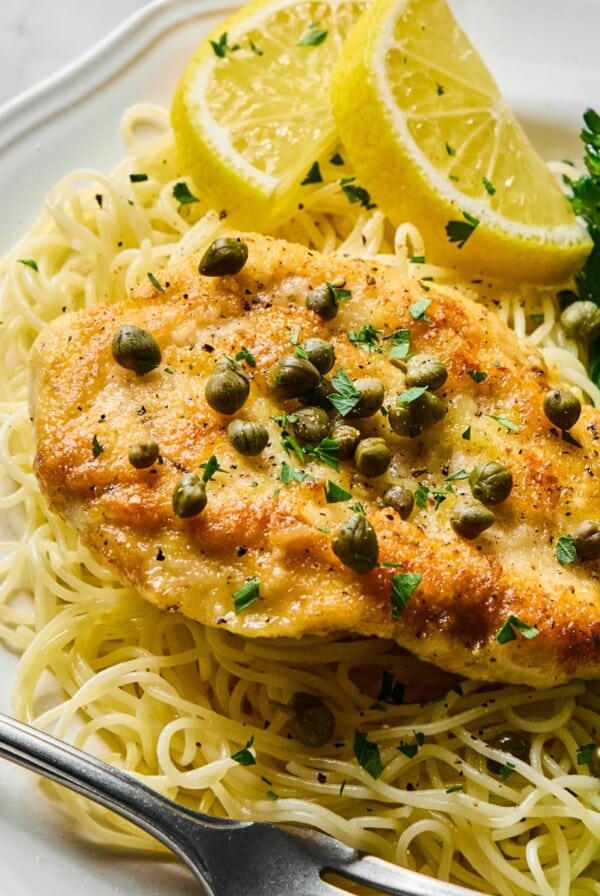 Healthy chicken piccata on a plate with pasta annd lemons on the side.