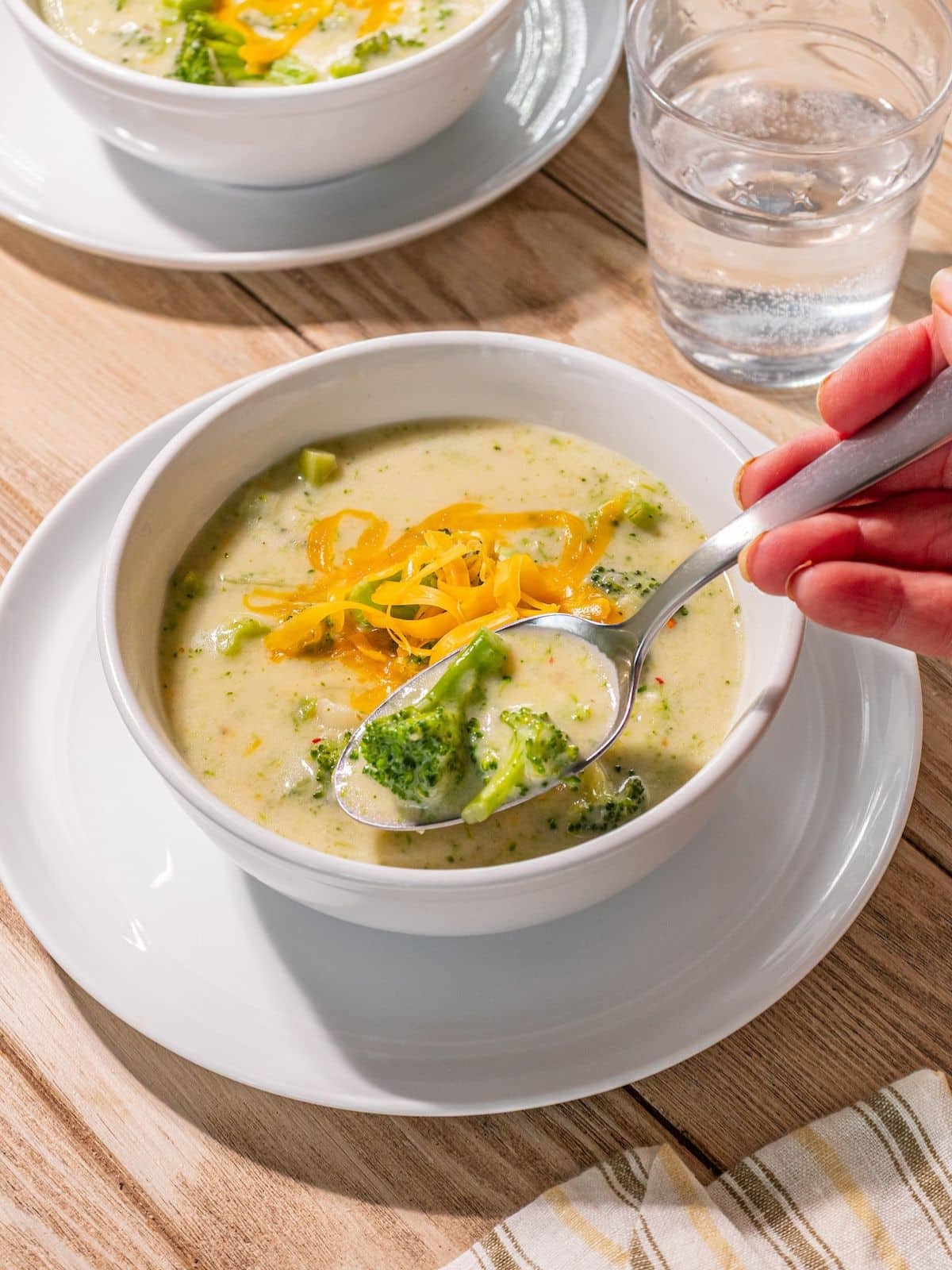 A spoon taking some broccoli soup with cheddar on top.