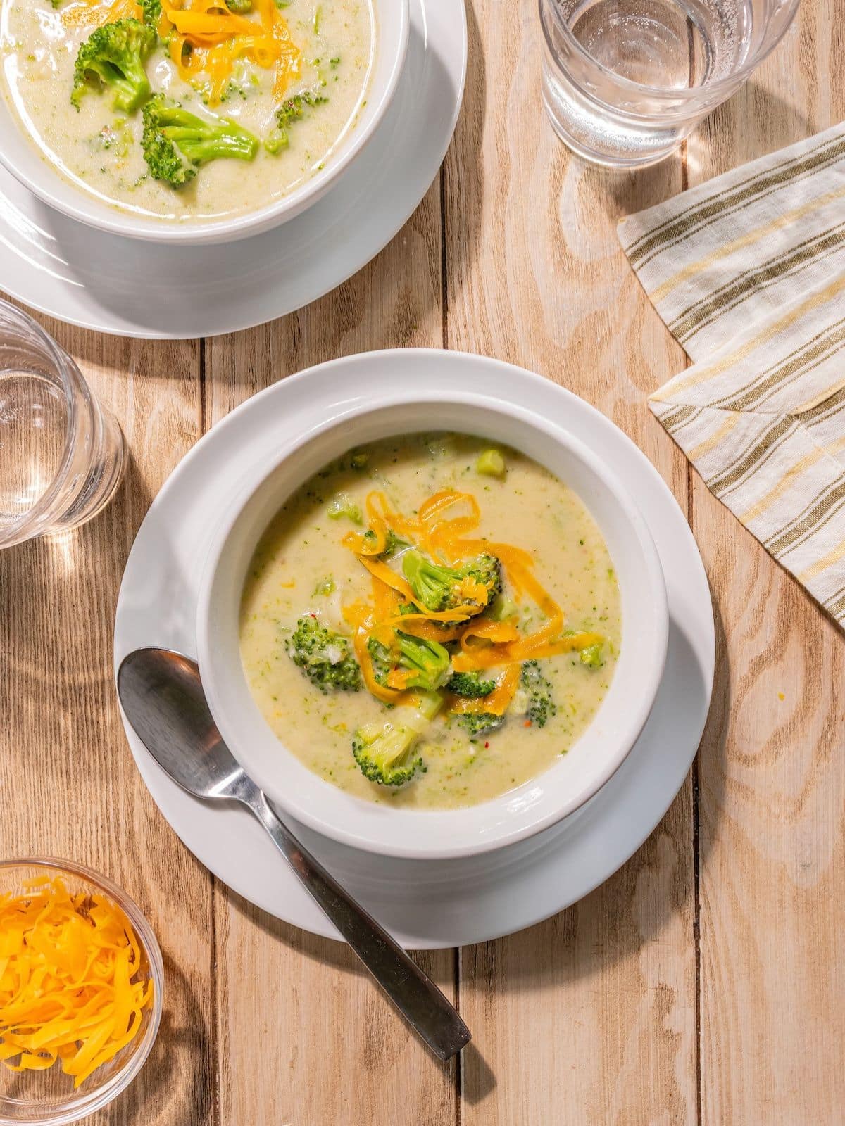 A bowl of broccoli soup with cheddar on top and a spoon on the side.