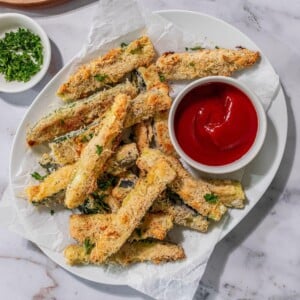 Baked zucchini sticks on a plate with ketchup on the side.